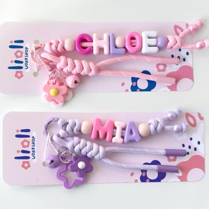 Peut inclure: Deux porte-cl&eacute;s nominatifs perl&eacute;s, l'un &eacute;pelant "CHLOE" en perles roses, violettes et blanches, et l'autre &eacute;pelant "MIA" en perles roses et violettes. Chaque porte-cl&eacute;s a un charme de fleur et un charme de cloche, pr&eacute;sent&eacute; sur une carte rose.