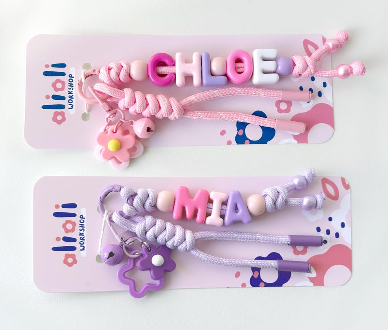Peut inclure: Deux porte-cl&eacute;s nominatifs perl&eacute;s, l'un &eacute;pelant "CHLOE" en perles roses, violettes et blanches, et l'autre &eacute;pelant "MIA" en perles roses et violettes. Chaque porte-cl&eacute;s a un charme de fleur et un charme de cloche, pr&eacute;sent&eacute; sur une carte rose.