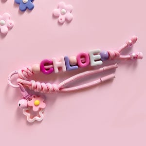 Peut inclure: Un porte-cl&eacute;s rose avec des perles &eacute;pelant le nom "CHLOE" avec des lettres color&eacute;es. Le porte-cl&eacute;s a un cordon rose et un charm en forme de fleur avec une clochette. Des d&eacute;corations en forme de fleurs sont &eacute;parpill&eacute;es.