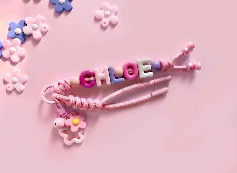 Peut inclure: Un porte-cl&eacute;s rose avec des perles &eacute;pelant le nom "CHLOE" avec des lettres color&eacute;es. Le porte-cl&eacute;s a un cordon rose et un charm en forme de fleur avec une clochette. Des d&eacute;corations en forme de fleurs sont &eacute;parpill&eacute;es.