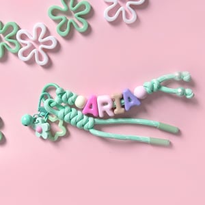 Peut inclure: Un porte-cl&eacute;s personnalis&eacute; avec le nom "MARIA" &eacute;pel&eacute; avec des perles de lettres color&eacute;es. Le porte-cl&eacute;s est orn&eacute; d'un cordon vert menthe, d'un charm en forme de fleur et de perles d&eacute;coratives. Le fond est rose clair avec des d&eacute;corations en forme de fleur.