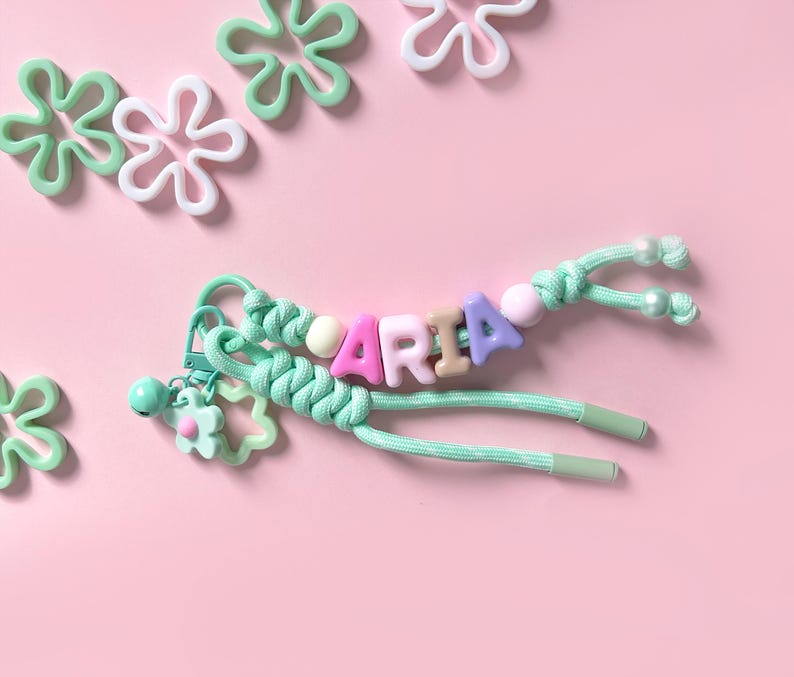 Peut inclure: Un porte-cl&eacute;s personnalis&eacute; avec le nom "MARIA" &eacute;pel&eacute; avec des perles de lettres color&eacute;es. Le porte-cl&eacute;s est orn&eacute; d'un cordon vert menthe, d'un charm en forme de fleur et de perles d&eacute;coratives. Le fond est rose clair avec des d&eacute;corations en forme de fleur.