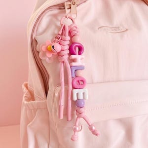Peut inclure: Un sac &agrave; dos rose clair avec un porte-cl&eacute;s rose assorti. Le porte-cl&eacute;s comporte un charm en forme de fleur, une cloche et des perles &eacute;pelant "CHLOE" en rose, violet et blanc. Le porte-cl&eacute;s est attach&eacute; &agrave; la fermeture &eacute;clair du sac &agrave; dos.