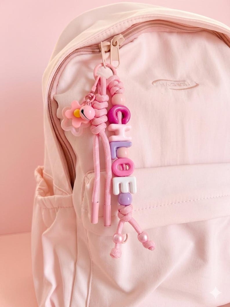 Peut inclure: Un sac &agrave; dos rose clair avec un porte-cl&eacute;s rose assorti. Le porte-cl&eacute;s comporte un charm en forme de fleur, une cloche et des perles &eacute;pelant "CHLOE" en rose, violet et blanc. Le porte-cl&eacute;s est attach&eacute; &agrave; la fermeture &eacute;clair du sac &agrave; dos.