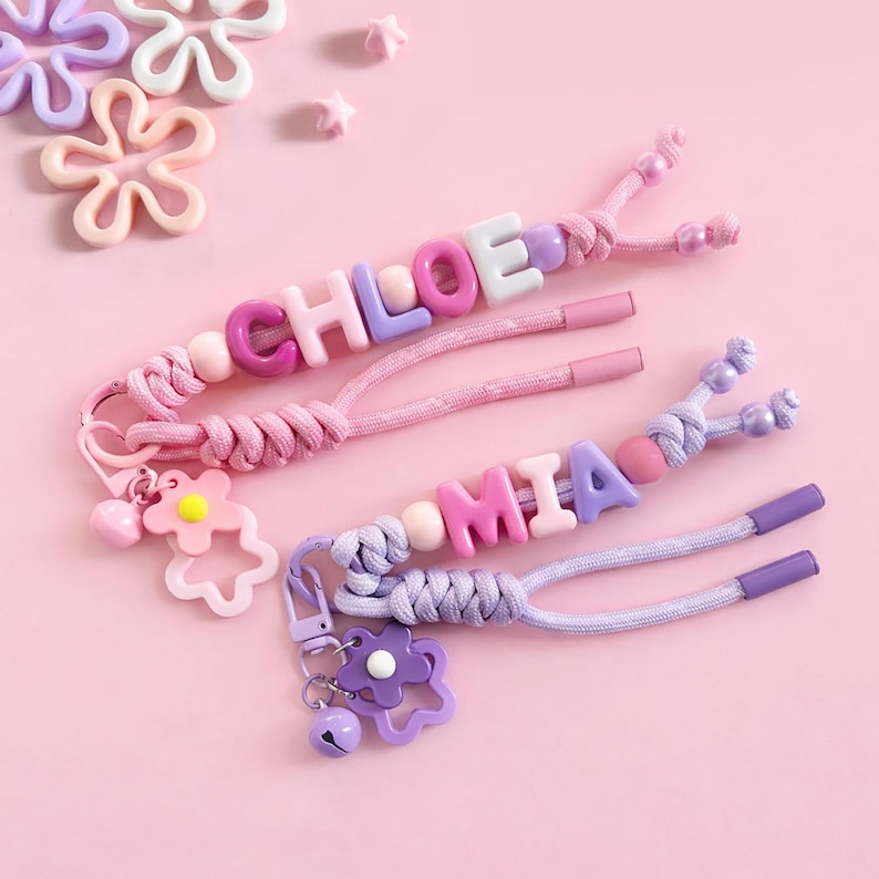 Peut inclure: Deux porte-cl&eacute;s personnalis&eacute;s avec les noms "CHLOE" et "MIA" &eacute;pel&eacute;s avec des perles de lettres color&eacute;es. Les porte-cl&eacute;s comportent des cordons roses et violets, des breloques en forme de fleurs et de petites cloches. Le fond est rose uni.