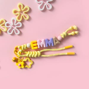 Peut inclure: Un porte-cl&eacute;s jaune avec le nom "EMMA" &eacute;pel&eacute; avec des perles de lettres color&eacute;es. Le porte-cl&eacute;s a un charme de fleur et est pos&eacute; sur un fond rose avec des d&eacute;corations en forme de fleurs &eacute;parpill&eacute;es.