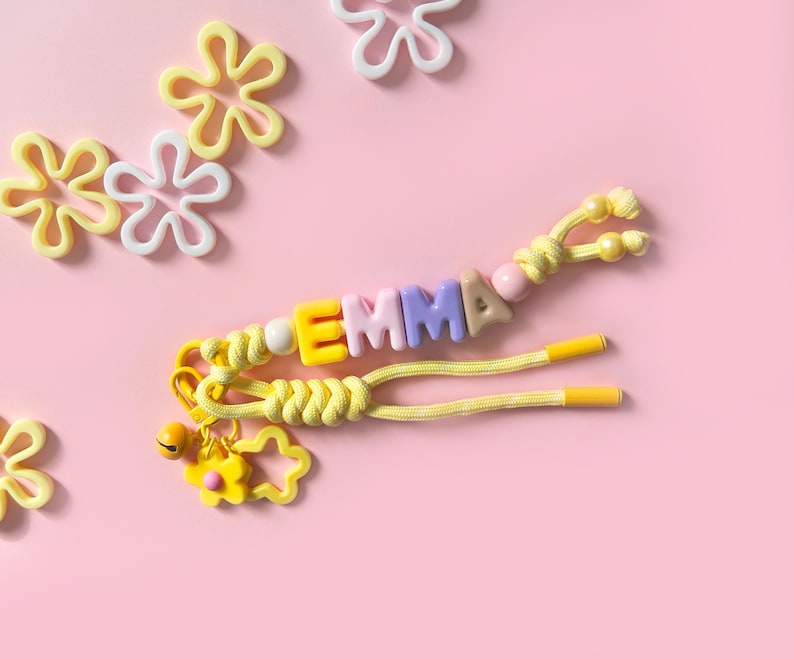 Peut inclure: Un porte-cl&eacute;s jaune avec le nom "EMMA" &eacute;pel&eacute; avec des perles de lettres color&eacute;es. Le porte-cl&eacute;s a un charme de fleur et est pos&eacute; sur un fond rose avec des d&eacute;corations en forme de fleurs &eacute;parpill&eacute;es.