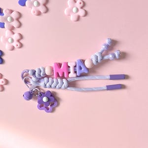 Peut inclure: Un porte-cl&eacute;s personnalis&eacute; avec le nom "MIA" &eacute;pel&eacute; avec des perles lettres roses et violettes. Le porte-cl&eacute;s comprend un cordon tress&eacute;, un charm en forme de fleur et une petite cloche. Des d&eacute;corations en forme de fleurs roses et violettes sont &eacute;parpill&eacute;es sur un fond rose.