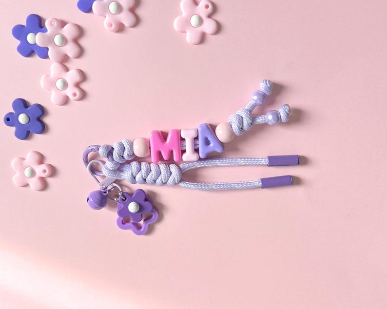 Peut inclure: Un porte-cl&eacute;s personnalis&eacute; avec le nom "MIA" &eacute;pel&eacute; avec des perles lettres roses et violettes. Le porte-cl&eacute;s comprend un cordon tress&eacute;, un charm en forme de fleur et une petite cloche. Des d&eacute;corations en forme de fleurs roses et violettes sont &eacute;parpill&eacute;es sur un fond rose.