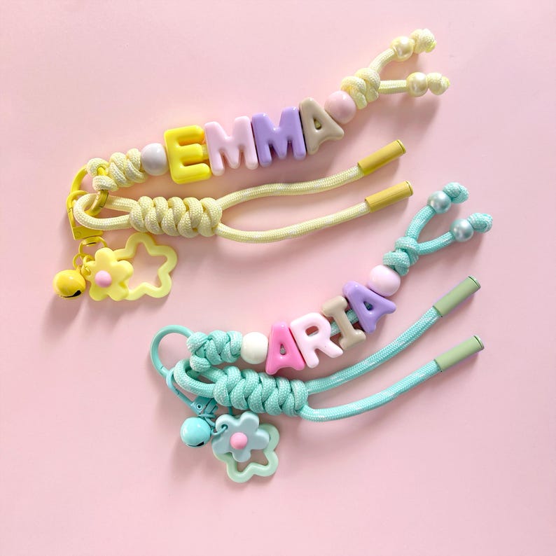 Peut inclure: Deux cordons personnalis&eacute;s avec des perles portant les noms "EMMA" et "ARIA" &eacute;pel&eacute;s avec des perles de lettres color&eacute;es. Les cordons sont jaunes et turquoise, avec des breloques en forme de fleur et d'&eacute;toile. Le fond est rose clair.