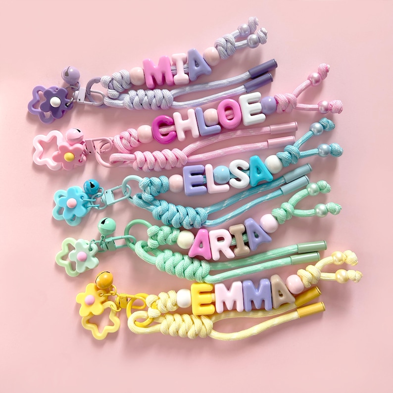 Peut inclure: Plusieurs porte-cl&eacute;s faits &agrave; la main avec des noms &eacute;crits en perles alphab&eacute;tiques. Les noms incluent MIA, CHLOE, ELSA, ARIA et EMMA. Les porte-cl&eacute;s sont de couleurs pastel et ont des breloques en forme de fleurs et de cloches.