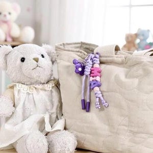 Peut inclure: Un ours en peluche blanc portant une robe blanche avec un col en dentelle est assis &agrave; c&ocirc;t&eacute; d'un sac fourre-tout beige matelass&eacute;. Un porte-cl&eacute;s perl&eacute; avec le nom "MIA" en rose et violet est suspendu au sac. D'autres ours en peluche sont visibles en arri&egrave;re-plan.