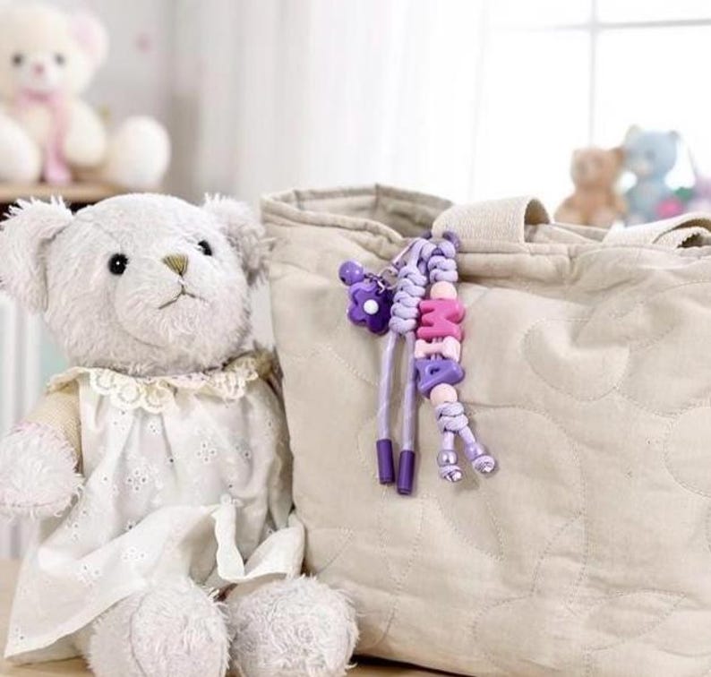 Peut inclure: Un ours en peluche blanc portant une robe blanche avec un col en dentelle est assis &agrave; c&ocirc;t&eacute; d'un sac fourre-tout beige matelass&eacute;. Un porte-cl&eacute;s perl&eacute; avec le nom "MIA" en rose et violet est suspendu au sac. D'autres ours en peluche sont visibles en arri&egrave;re-plan.