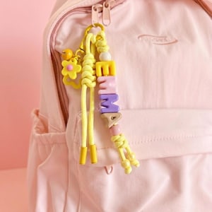 Peut inclure: Un sac &agrave; dos rose clair avec un porte-cl&eacute;s jaune. Le porte-cl&eacute;s &eacute;pelle "EMMA" avec des perles color&eacute;es. Un charm en forme de fleur jaune et des cordons jaunes sont &eacute;galement attach&eacute;s au porte-cl&eacute;s.