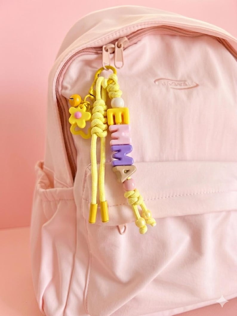 Peut inclure: Un sac &agrave; dos rose clair avec un porte-cl&eacute;s jaune. Le porte-cl&eacute;s &eacute;pelle "EMMA" avec des perles color&eacute;es. Un charm en forme de fleur jaune et des cordons jaunes sont &eacute;galement attach&eacute;s au porte-cl&eacute;s.
