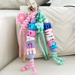 Peut inclure: Porte-cl&eacute;s personnalis&eacute;s roses et vert menthe, &eacute;pelant les noms "GRACE" et "EMMA" avec des perles de lettres color&eacute;es. Chaque porte-cl&eacute;s comporte des accents de fleurs et de perles, attach&eacute;s &agrave; un sac &agrave; main blanc.