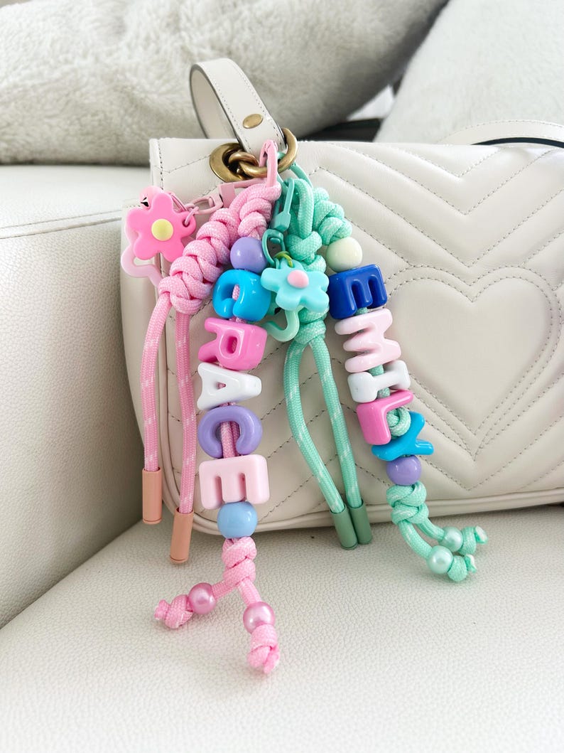 Peut inclure: Porte-cl&eacute;s personnalis&eacute;s roses et vert menthe, &eacute;pelant les noms "GRACE" et "EMMA" avec des perles de lettres color&eacute;es. Chaque porte-cl&eacute;s comporte des accents de fleurs et de perles, attach&eacute;s &agrave; un sac &agrave; main blanc.