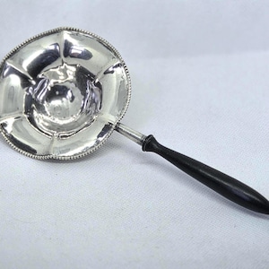 May include: A silver punch ladle featuring a wide, circular, scalloped bowl with a beaded rim and a smooth, tapered black wooden handle that terminates in a pointed finial. The reflective metallic surface shows a polished, bright finish.