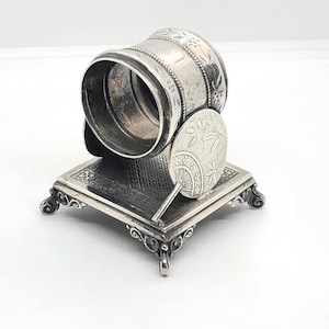 May include: An antique silver napkin ring with a decorative fan and a square base. The napkin ring is cylindrical with floral engravings. The fan is round with a floral design. The base has ornate detailing and scrolled feet.