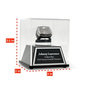 May include: A championship ring in a clear display case. The ring is silver with a diamond-studded top and the word "CHAMPION". The black base has the text "Johnny Lawrence Cobra Kai 2023 Fantasy Football Champion". The case is 8.9 cm high and 7.6 cm wide.
