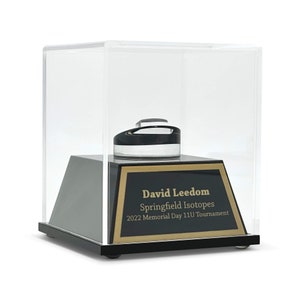 May include: A black and clear display case with a black base and a gold-trimmed plaque. The plaque reads "David Leedom Springfield Isotopes 2022 Memorial Day 11U Tournament". Inside the case is a black and clear object.