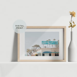 May include: A framed digital print featuring a coastal scene with a sign that reads "Solana Beach" in turquoise. The image is in a light wood frame, with a white vase holding a dried flower to the right.