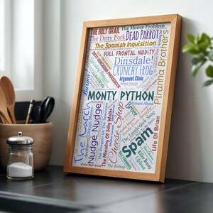 May include: A wooden framed print featuring the words "Monty Python" in a central position, surrounded by phrases and words in various colours and fonts, including "Spam" and "Crunchy Frog". The frame is light-colored.