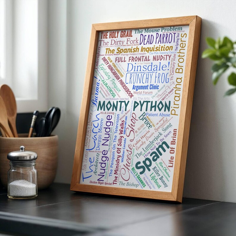 May include: A wooden framed print featuring the words "Monty Python" in a central position, surrounded by phrases and words in various colours and fonts, including "Spam" and "Crunchy Frog". The frame is light-colored.