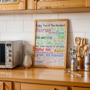 Puede incluir: Un cuadro con frases coloridas como "Bazinga!" y "How Sweet It Is!" se muestra en una encimera de madera. También se ven utensilios de cocina y un microondas. El cuadro mide aproximadamente 40 x 50 cm.