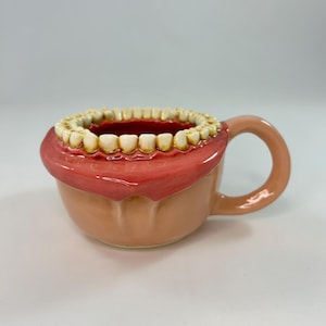 May include: A novelty ceramic mug shaped like a mouth, featuring a full set of teeth around the rim. The mug has a peach-colored base and handle, with a red interior and pink lips. A unique and quirky gift idea.