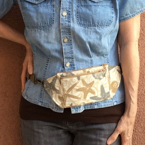 May include: A brown fabric fanny pack with a zipper closure and a white and brown starfish print. The fanny pack is worn over a denim shirt and brown pants.