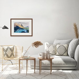 Pode incluir: Uma pintura de aquarela emoldurada de uma cordilheira com um lago em primeiro plano. A pintura est&aacute; pendurada em uma parede branca em um ambiente de sala de estar com um sof&aacute;, duas mesas laterais e uma cadeira.