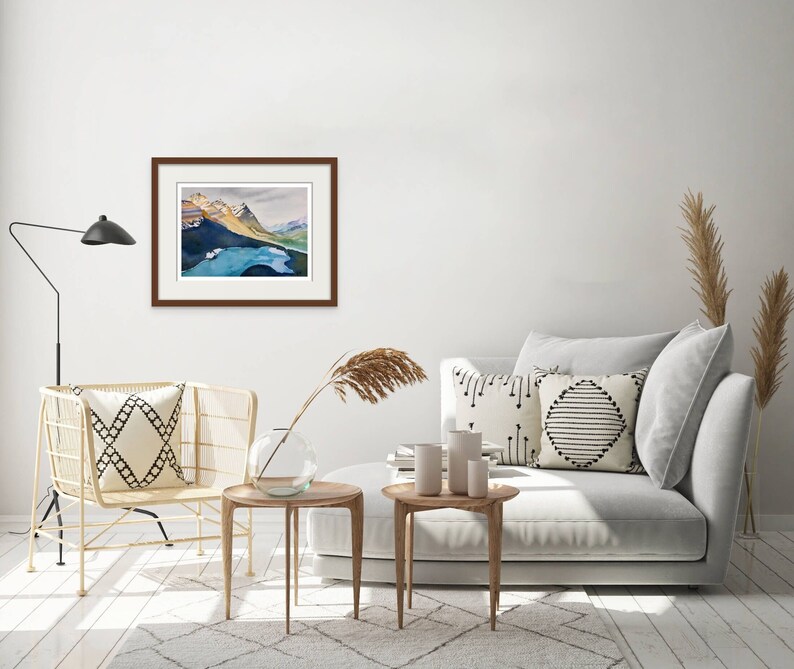 Pode incluir: Uma pintura de aquarela emoldurada de uma cordilheira com um lago em primeiro plano. A pintura est&aacute; pendurada em uma parede branca em um ambiente de sala de estar com um sof&aacute;, duas mesas laterais e uma cadeira.