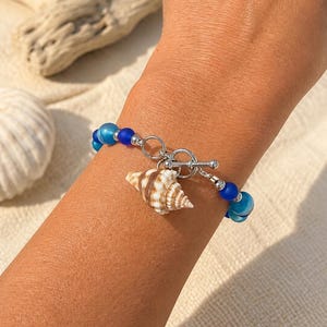 May include: A blue beaded bracelet with a seashell charm. The bracelet has a toggle clasp and is made with blue and clear beads. The seashell charm is brown and white.