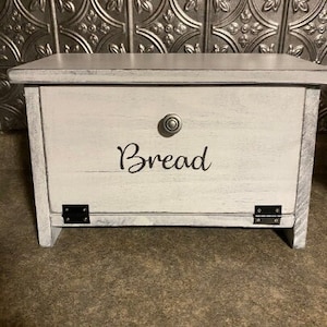 May include: A white wooden bread box with a distressed finish. The word "Bread" is written in a cursive font on the front. Features a knob and hinges. The box is sitting on a brown surface.
