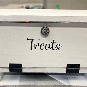 K&ouml;nnte beinhalten: Wei&szlig;e Holzkiste mit einem Scharnierdeckel und einem Metallknopf. Die Kiste hat einen schwarzen Vinyl-Aufkleber mit der Aufschrift "Treats".