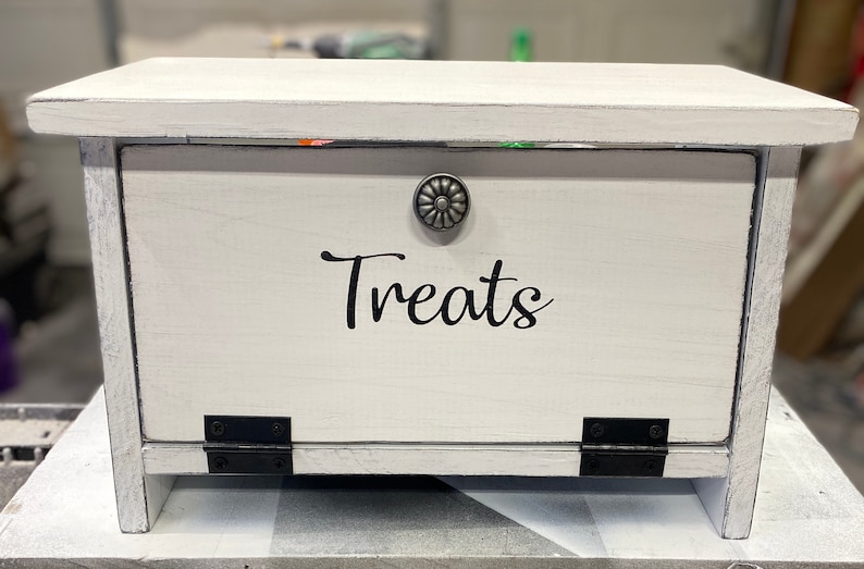 K&ouml;nnte beinhalten: Wei&szlig;e Holzkiste mit einem Scharnierdeckel und einem Metallknopf. Die Kiste hat einen schwarzen Vinyl-Aufkleber mit der Aufschrift "Treats".