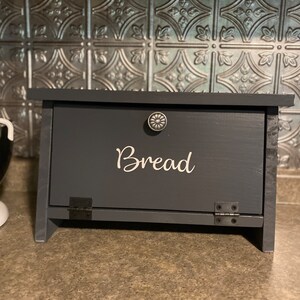 May include: A dark gray wooden bread box with a black metal hinge and a white inscription that reads "Bread".