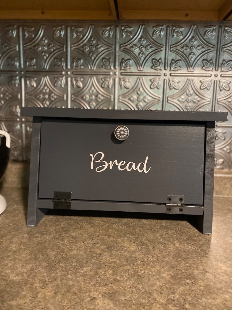 May include: A dark gray wooden bread box with a black metal hinge and a white inscription that reads "Bread".