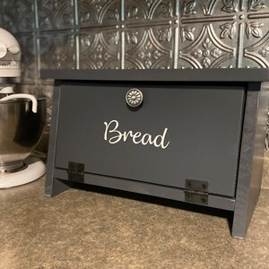 K&ouml;nnte beinhalten: Eine dunkelgraue Brotbox aus Holz mit einem Scharnierdeckel und einem silbernen Knopf. Die Box hat einen wei&szlig;en Vinyl-Aufkleber mit der Aufschrift "Brot".