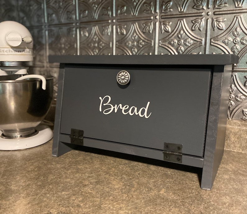 K&ouml;nnte beinhalten: Eine dunkelgraue Brotbox aus Holz mit einem Scharnierdeckel und einem silbernen Knopf. Die Box hat einen wei&szlig;en Vinyl-Aufkleber mit der Aufschrift "Brot".