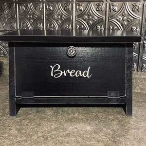K&ouml;nnte beinhalten: Schwarze Brotbox aus Holz mit einer wei&szlig;en Aufschrift "Brot". Die Box hat einen Klappdeckel und einen Metallknopf.