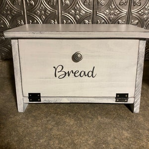 K&ouml;nnte beinhalten: Wei&szlig;er Brotbeh&auml;lter aus Holz mit einem schwarzen Metallscharnier und einem silbernen Knopf. Der Beh&auml;lter hat das Wort "Brot" in Schwarz auf der Vorderseite.