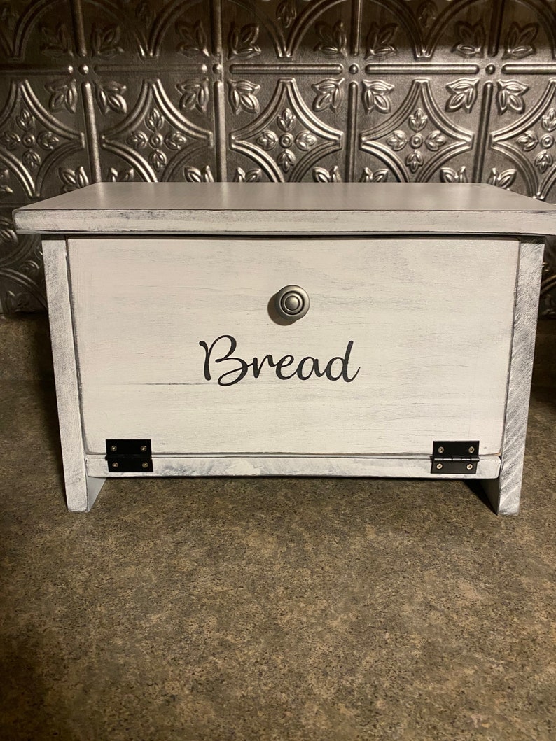 K&ouml;nnte beinhalten: Wei&szlig;er Brotbeh&auml;lter aus Holz mit einem schwarzen Metallscharnier und einem silbernen Knopf. Der Beh&auml;lter hat das Wort "Brot" in Schwarz auf der Vorderseite.