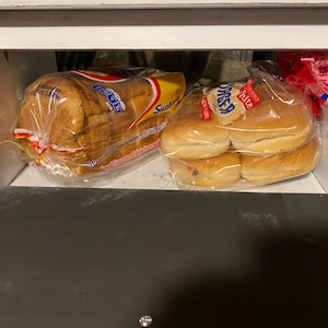 K&ouml;nnte beinhalten: Ein wei&szlig;es Brotlaib in einer durchsichtigen Plastikt&uuml;te mit einem roten und gelben Etikett, auf dem "Sandwich" steht, und eine T&uuml;te mit wei&szlig;en Hamburgerbr&ouml;tchen in einer durchsichtigen Plastikt&uuml;te mit einem roten und wei&szlig;en Etikett, auf dem "Kitte" steht, in einer wei&szlig;en Holzbrotbox.