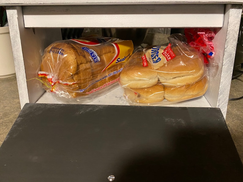 K&ouml;nnte beinhalten: Ein wei&szlig;es Brotlaib in einer durchsichtigen Plastikt&uuml;te mit einem roten und gelben Etikett, auf dem "Sandwich" steht, und eine T&uuml;te mit wei&szlig;en Hamburgerbr&ouml;tchen in einer durchsichtigen Plastikt&uuml;te mit einem roten und wei&szlig;en Etikett, auf dem "Kitte" steht, in einer wei&szlig;en Holzbrotbox.