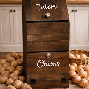 May include: A dark brown wooden storage bin with two compartments labeled "Taters" and "Onions" in white script. The bin has a hinged top and a drawer, each with a decorative knob. Potatoes and onions are visible.
