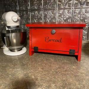 K&ouml;nnte beinhalten: Rote Holzbrotbox mit schwarzem Metallgriff und Scharnieren. Die Box hat das Wort "Brot" in schwarzen Buchstaben auf der Vorderseite.