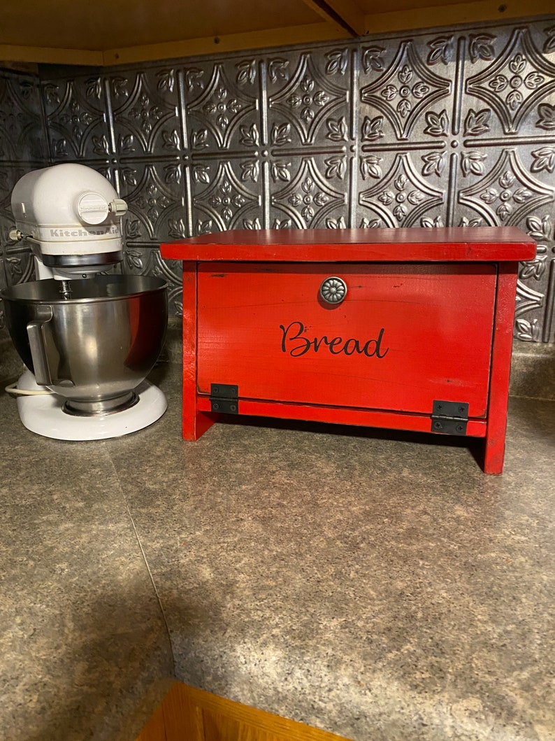 K&ouml;nnte beinhalten: Rote Holzbrotbox mit schwarzem Metallgriff und Scharnieren. Die Box hat das Wort "Brot" in schwarzen Buchstaben auf der Vorderseite.
