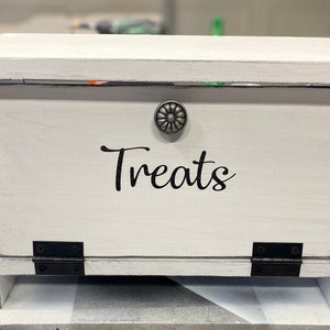 May include: White wooden box with a hinged lid and a silver knob. The box has a black label that says "Treats".